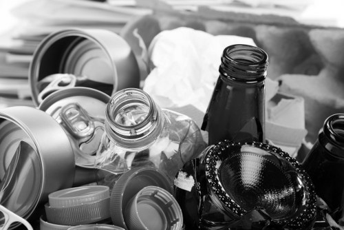 A collection of various types of discarded rubbish, including open glass bottles, aluminum cans with pull-tab openings, plastic bottles with caps, and metal or plastic containers. The bottles are scattered and partly leaning against each other, with some lying on their sides, in an outdoor setting on a flat surface, possibly a pavement or driveway. Surrounding the rubbish are crumpled paper or tissue, indicating clutter. In the background, blurred objects suggest an area where waste has accumulated, with no specific environmental features visible. The image conveys a scene suitable for waste removal services, such as those offered by Office Clearance Highgate, focusing on the types of waste typical during clearance or rubbish collection in a London suburb.