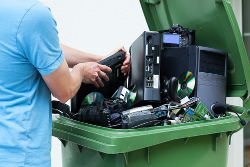 A person wearing a light blue t-shirt is standing outdoors next to a large green waste disposal bin filled with discarded electronic waste and broken household appliances. Visible items include an old desktop computer tower with a black casing, several CDs, a digital camera, and various cables and circuitry, all jumbled together inside the bin. The background suggests an urban or suburban environment, possibly on a driveway or pavement. The scene reflects a professional rubbish removal service, such as that provided by Office Clearance Highgate, which handles the disposal of electronic waste and household debris. The lighting appears natural, highlighting the textures of the electronic components and the smooth, matte surface of the bin. This image underscores the importance of responsible rubbish disposal in the Highgate area, supporting environmentally compliant waste clearance for local clients looking to declutter residential or commercial spaces.