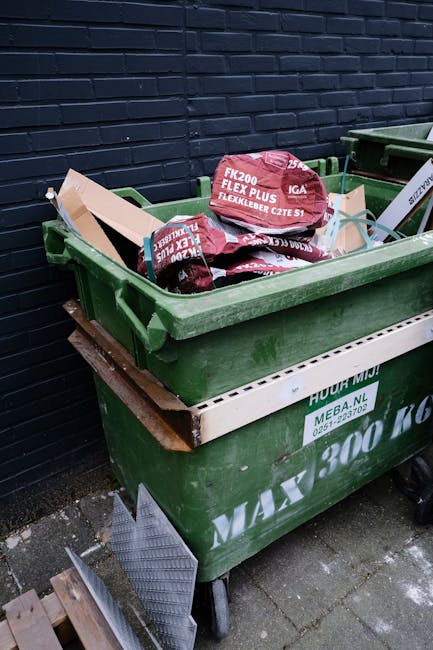 Two green plastic rubbish bins with hinged lids are positioned side by side on a concrete pavement at the edge of a street, adjacent to a curb. The bins appear to be made of durable, smooth plastic wi