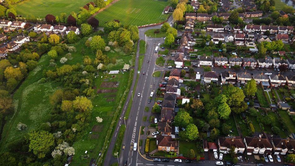 An aerial view of a dense residential area showing clustered houses with pitched roofs in various shades of grey, brown, and red. The properties are separated by narrow driveways and small green garde