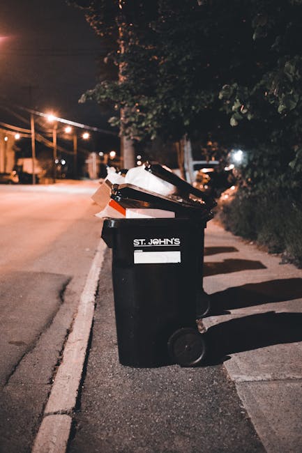 A large outdoor area filled with accumulated waste and recycling materials, including multiple stacks of flattened cardboard boxes with colorful fruit and fresh produce branding, arranged in front of
