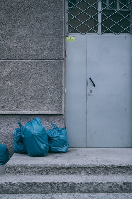 A collection of black rubbish bags is piled up directly in front of a red metal door mounted within a textured, off-white building wall. The bags appear to contain household waste and are stacked haph