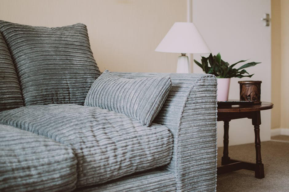 A close-up view of a light grey, ribbed fabric sofa situated in a living room, with a matching cushion resting on the seat. Behind the sofa, there is a cream-colored wall and a white table lamp with a