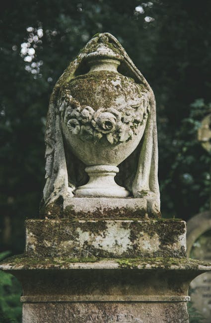 An aged stone urn with a weathered surface and moss growth, positioned on a rectangular stone pedestal in an outdoor setting. The urn features a rounded body with a decorative floral relief carved ont