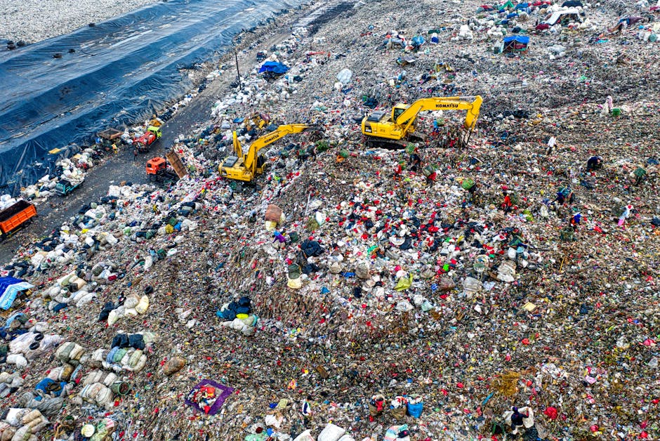 A large mound of assorted scrap metal and discarded electronic waste is piled outdoors under a clear blue sky. The heap includes twisted steel, bent metal panels, damaged appliances, and fragmented el
